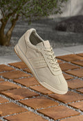 Beige Tommy Jeans Sneaker mit Schnürsenkeln, gepolsterter Sohle und markierten Seitenstreifen, schwebt über einem Boden aus Holz- und Steinfliesen im Freien.