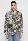 Junger Mann trägt eine hochgeschlossene Zip-Jacke mit einem abstrakten Muster in Grau, Weiß, Gelb und Grün sowie eine Bluejeans.