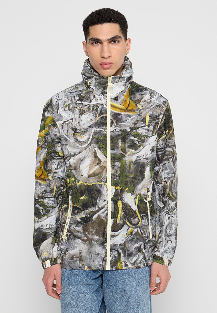 Junger Mann trägt eine hochgeschlossene Zip-Jacke mit einem abstrakten Muster in Grau, Weiß, Gelb und Grün sowie eine Bluejeans.