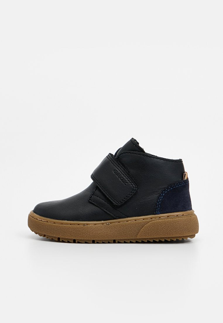 Schwarze Leder-Stiefelette mit einem Klettverschluss, beigefarbener Gummisohle und blauen Wildlederakzenten an der Ferse. Glatte Textur mit runder Zehenform.