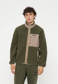 Junger Mann trägt eine grüne Fleecejacke mit beiger Brusttasche und Reißverschluss, kombiniert mit olivgrünen Hosen mit Text am linken Oberschenkel.