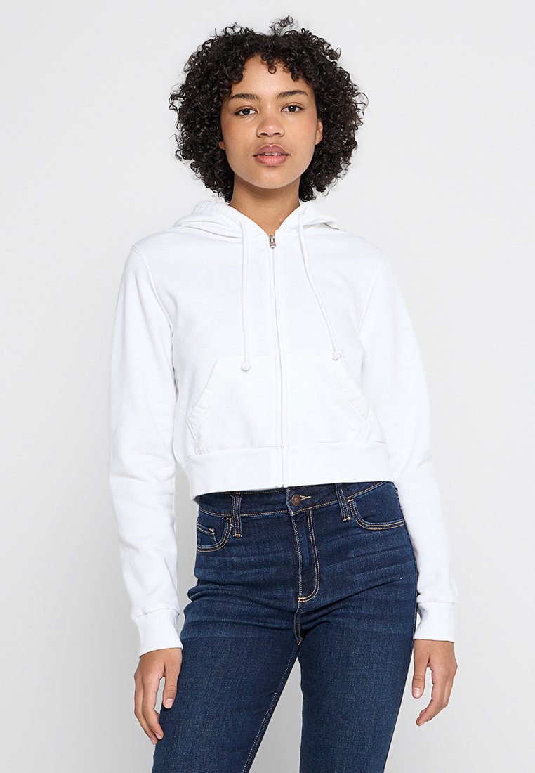 Gekürzte weiße Zip-Hoodie mit Kordeln, Fronttaschen und gerippten Bündchen. Kombiniert mit dunkelblauen Denim-Jeans.
