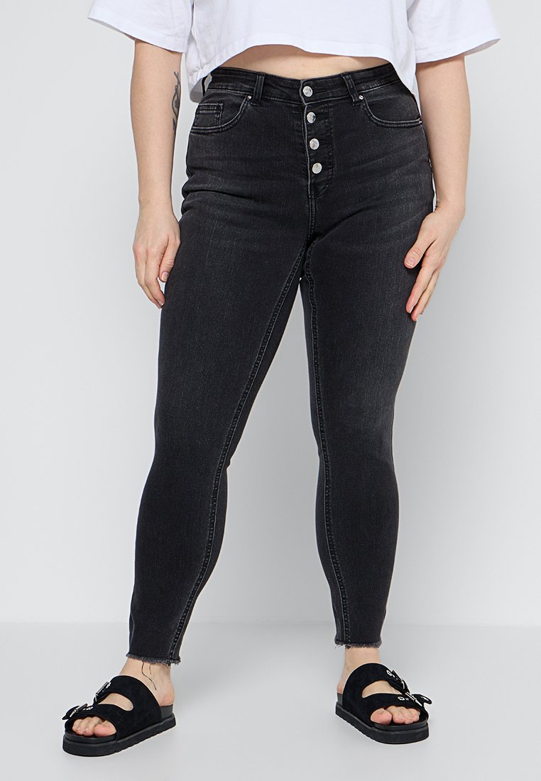 Hoch taillierte schwarze Jeans aus Denim mit schmalem Schnitt, fünf Knöpfen,_fronttaschen und fransigem Saum, kombiniert mit schwarzen Schlüpfschuhen.