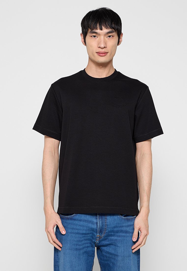 Schwarzes Baumwoll-T-Shirt mit Rundhalsausschnitt und kurzen Ärmeln, mit einem dezenten Logodetail auf der Brust. Kombiniert mit blauen Jeans aus Denim.