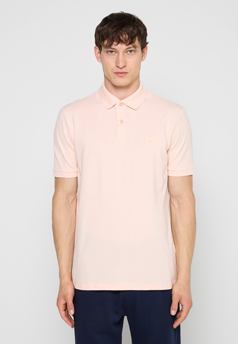 Mann trägt ein hellrosa kurzärmeliges Poloshirt und dunkle Hose, steht vor einem schlichten hellen Hintergrund.