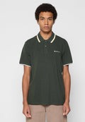 Mann trägt ein dunkelgrünes Poloshirt mit hellfarbiger Einfassung und beigen Hosen, steht vor einem schlichten hellgrauen Hintergrund.