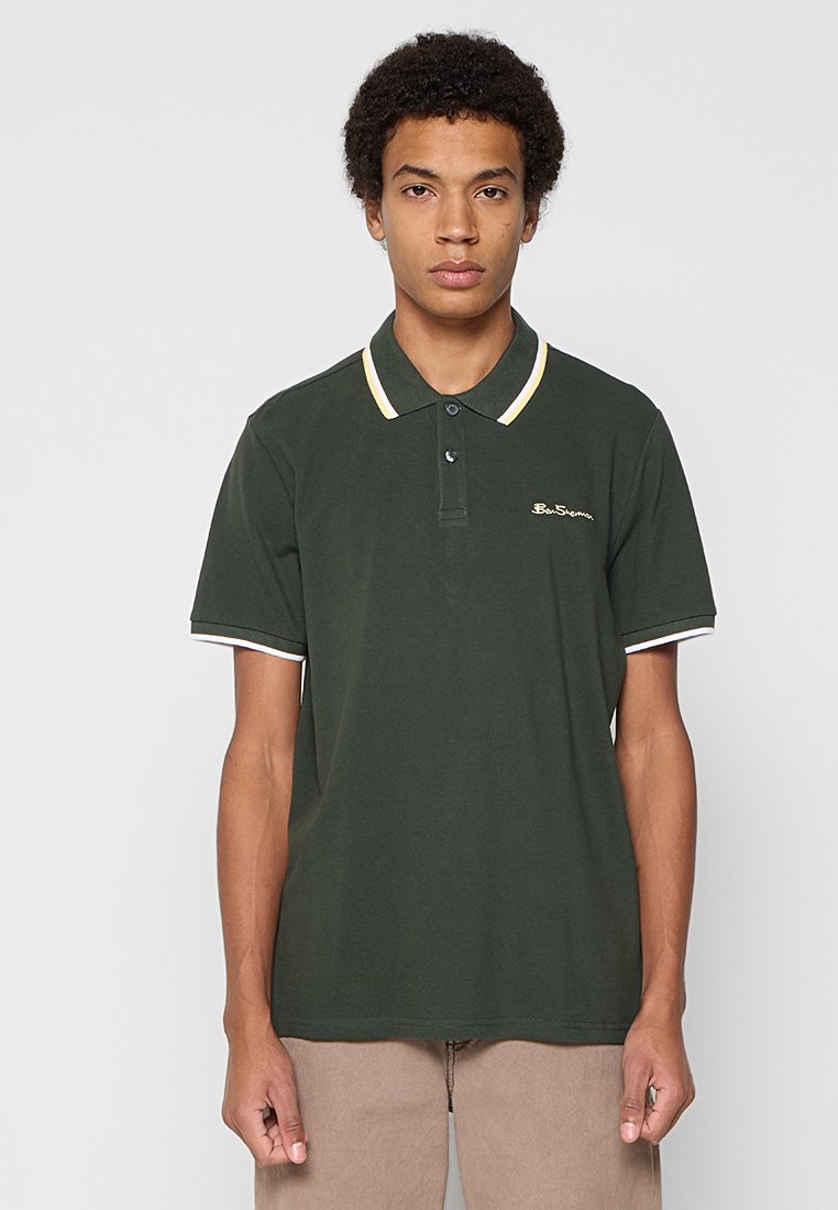 Mann trägt ein dunkelgrünes Poloshirt mit hellfarbiger Einfassung und beigen Hosen, steht vor einem schlichten hellgrauen Hintergrund.