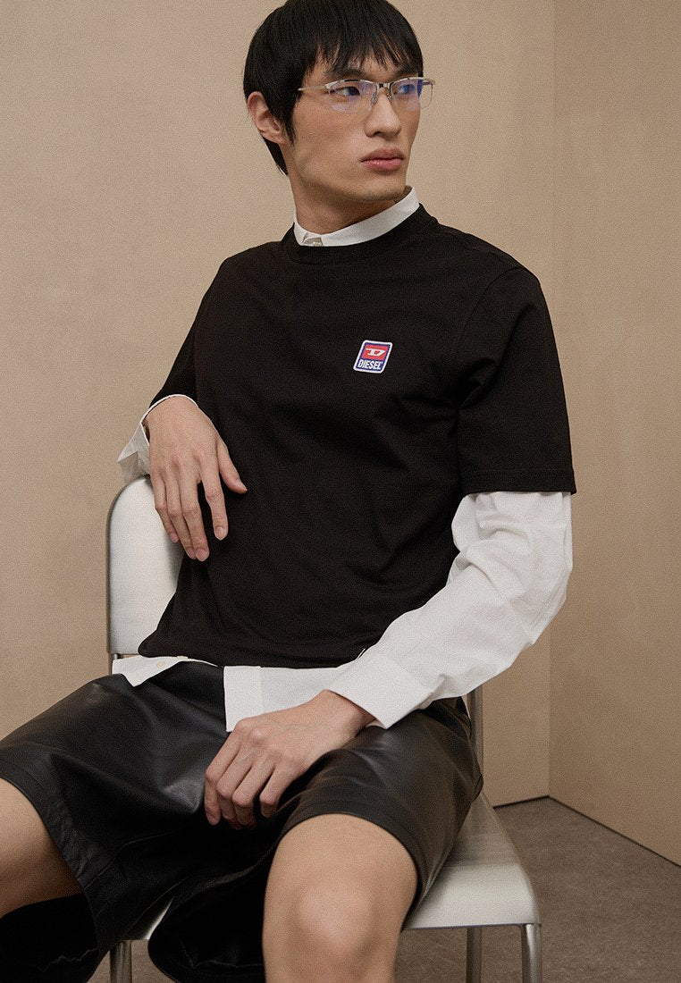 Schwarzes Baumwoll-T-Shirt mit rundem Ausschnitt, versehen mit einem kleinen, bunten "Diesel"-Logo. Wird über einem weißen Langarmshirt getragen und mit schwarzen Shorts kombiniert.