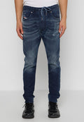 Mann in dunkelblauen, ausgewaschenen Jeans mit Zugband, schwarzen Sneakers und einem weißen Hemd, der vor einem schlichten Hintergrund steht.