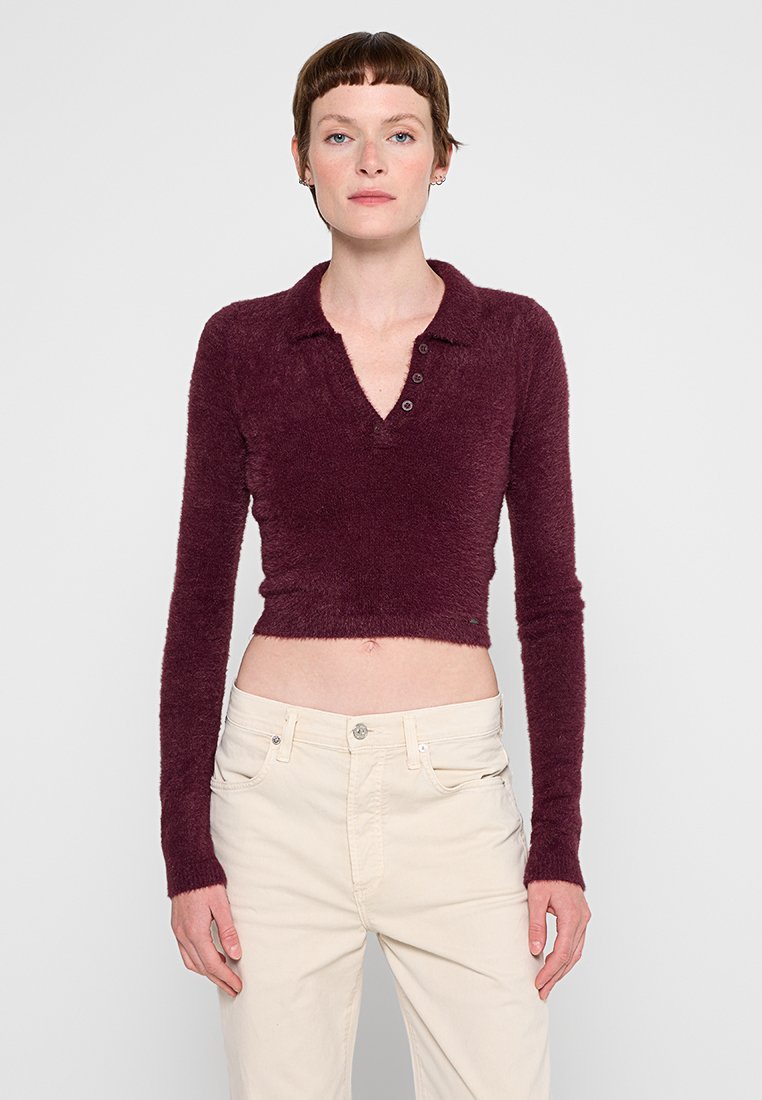 Burgunderfarbiges, cropped Langarm-Polohemd aus weichem, fuzzy Material mit einer Drei-Button-Leiste und einem Kragen. Getragen mit hellen Hosen.