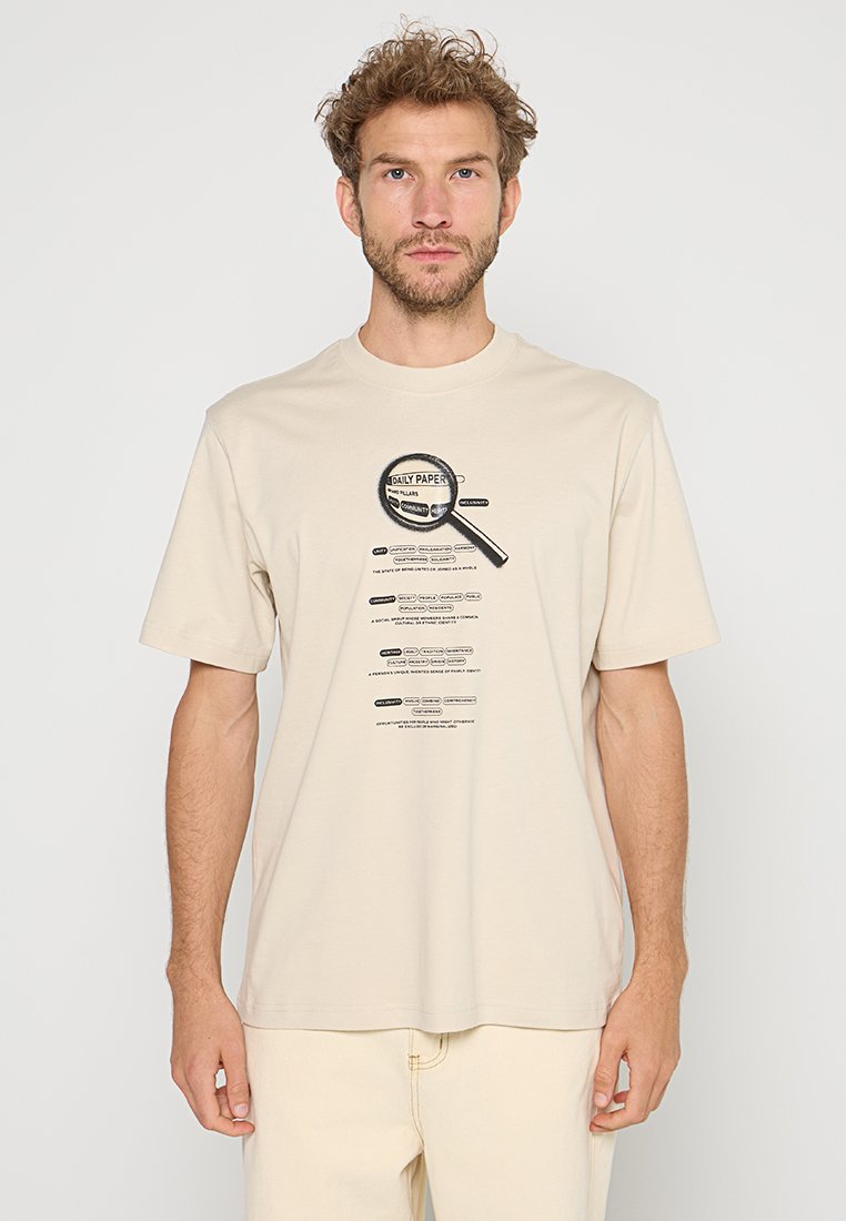 Mann mit lockigem Haar trägt ein beigefarbenes T-Shirt mit einem schwarzen Vergrößerungsglas-Grafik und kleinem Text darunter.
