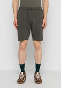 Mann trägt dunkelgrüne Shorts mit Kordelzug, dunkelgrüne Socken und braune Sneaker und steht vor einem einfarbigen Hintergrund.