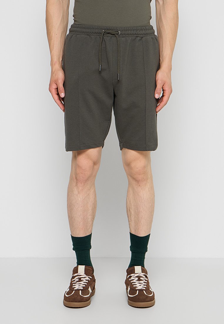 Mann trägt dunkelgrüne Shorts mit Kordelzug, dunkelgrüne Socken und braune Sneaker und steht vor einem einfarbigen Hintergrund.