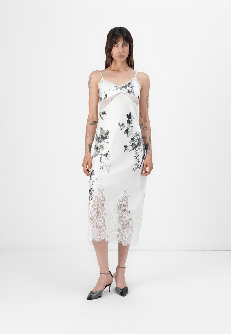 Satin-Nachtkleid mit floralem Muster, weißem Grund, Spitzenbesatz am Saum, verstellbaren Spaghettiträgern und einem lockeren Schnitt. Dazu werden schwarze Absätze getragen.
