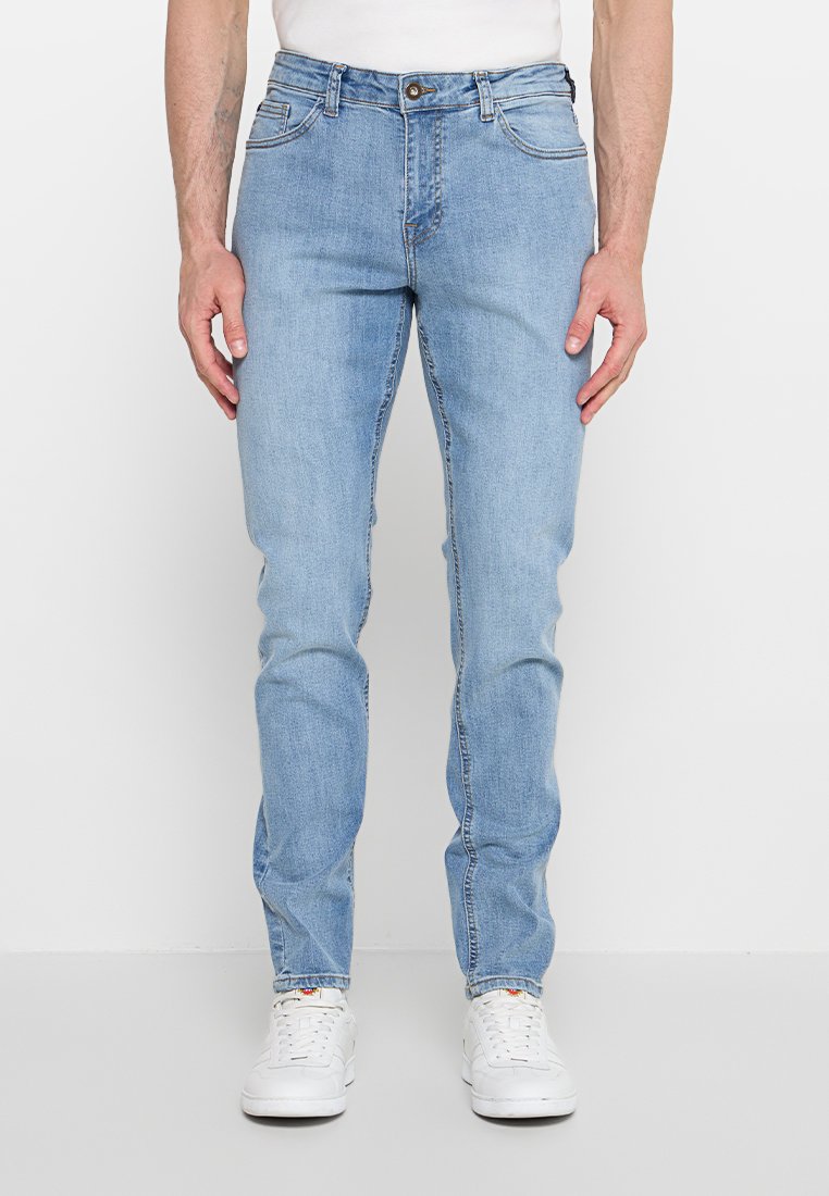 Mann trägt hellblaue Skinny-Jeans und weiße Sneakers, steht vor einem schlichten weißen Hintergrund.
