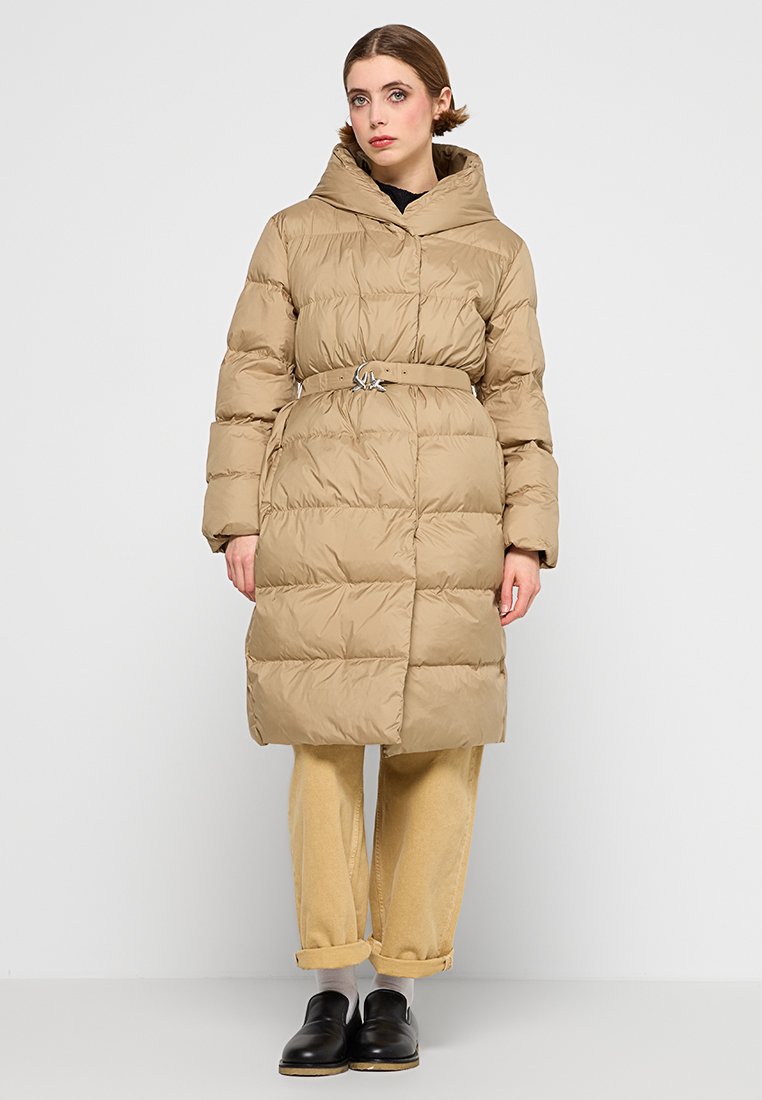 Beige Pufferjacke mit gerafftem Gürtel und silberner Schnalle, ausgestattet mit einem hohen Kragen, langen Ärmeln und steppender Textur. Getragen über hellen Hosen.