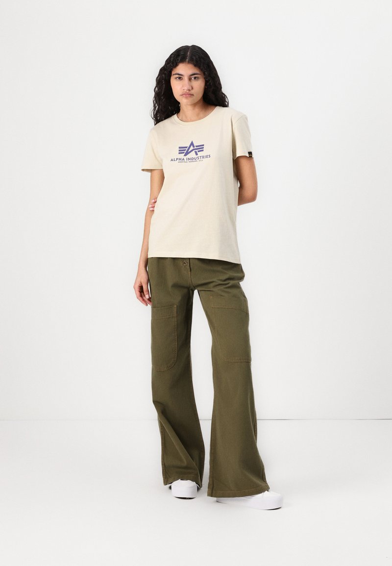 Junge Frau mit langen lockigen Haaren trägt ein beigefarbenes Alpha Industries T-Shirt, olivgrüne weite Hosen und weiße Turnschuhe, steht vor einer weißen Wand.