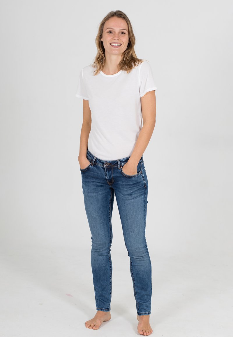 Weißes kurzärmeliges T-Shirt kombiniert mit blauen Skinny-Jeans. Der Stoff wirkt glatt mit einer klassischen Passform. Barfußhaltung vor einem schlichten Hintergrund.