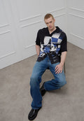 Schwarzes Kurzarmhemd mit blau-weißem abstraktem Muster, kombiniert mit hellblauen Jeans und schwarzen Schuhen. Modell sitzt auf einem Stuhl.