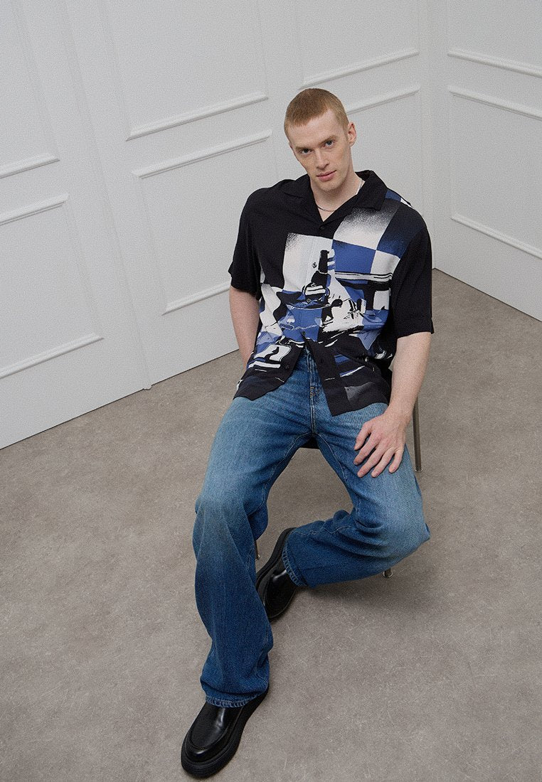 Schwarzes Kurzarmhemd mit blau-weißem abstraktem Muster, kombiniert mit hellblauen Jeans und schwarzen Schuhen. Modell sitzt auf einem Stuhl.