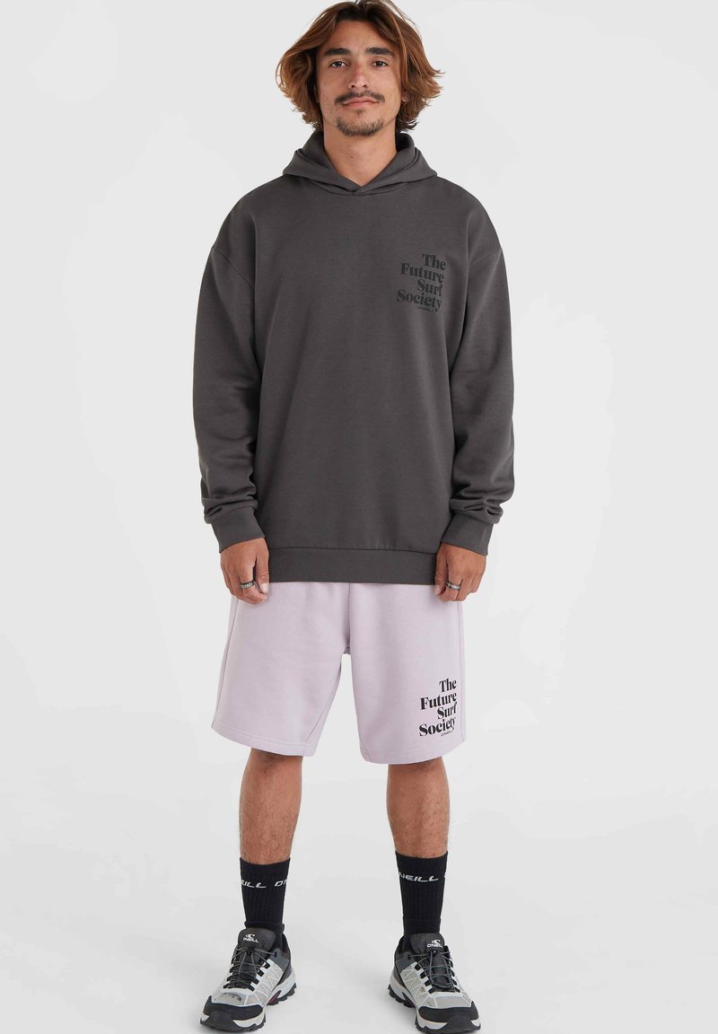 Grauer Hoodie mit schwarzem Logo auf der Brust, kombiniert mit hellrosa Shorts, die ein ähnliches Logo haben. Schwarze Socken und graue Sneakers runden den Look ab.