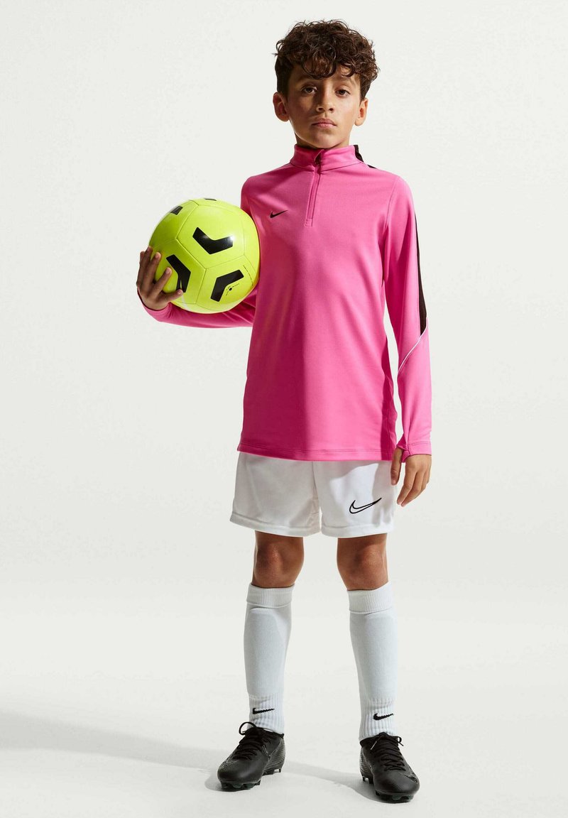Junge in pinkem langärmligen Sporttrikot, weißen Shorts und schwarzen Fußballschuhen, der einen gelb-schwarzen Fußball hält und vor einem einfarbigen Hintergrund steht.