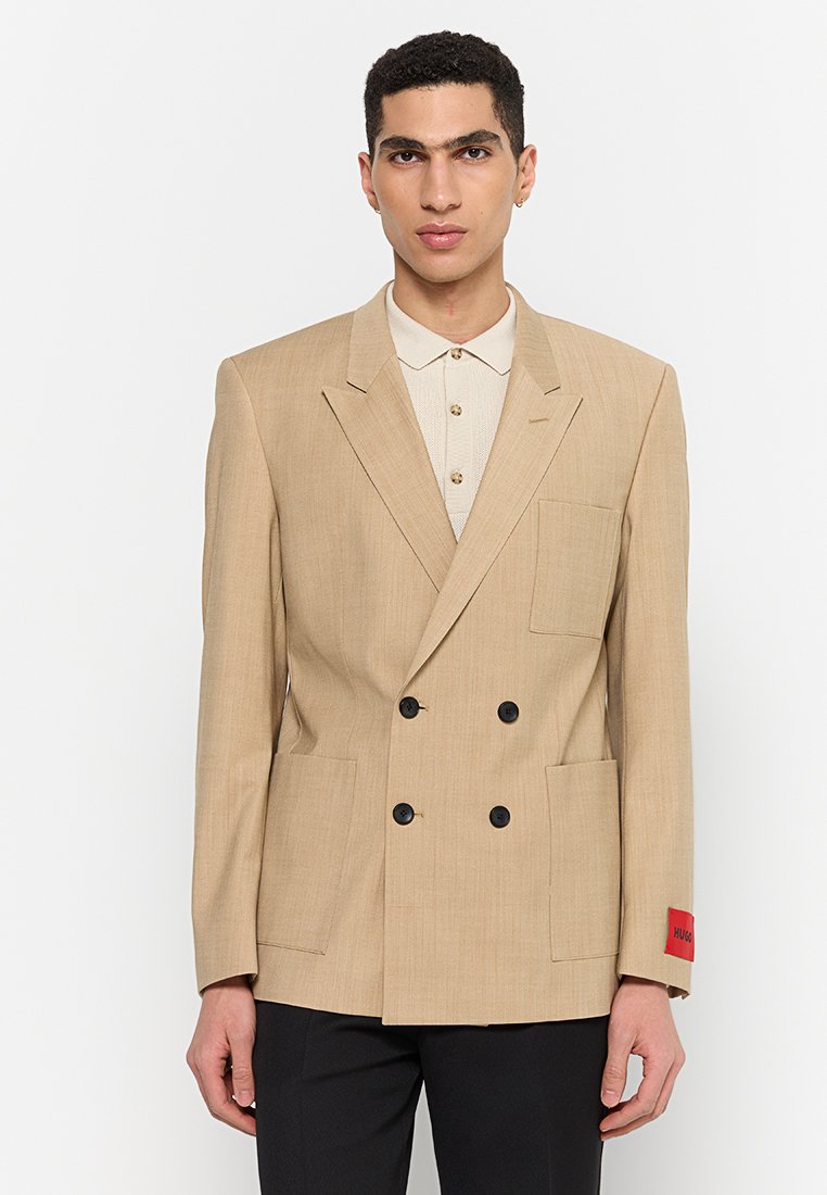 Beige, doppelt geknöpfter Blazer mit zwei großen Taschen, schwarzen Knöpfen und einem roten Etikett am Ärmel. Strukturierte, maßgeschneiderte Gestaltung und klar definierte Form.