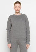 Junge Person mit heller Haut und nach hinten gebundenem Haar, die einen grauen Kapuzenpullover und passende Jogginghosen mit "Peak Performance"-Logos trägt.