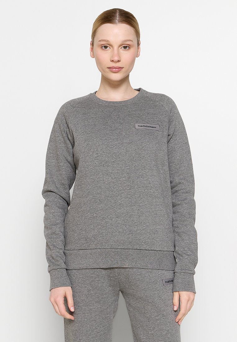 Junge Person mit heller Haut und nach hinten gebundenem Haar, die einen grauen Kapuzenpullover und passende Jogginghosen mit "Peak Performance"-Logos trägt.