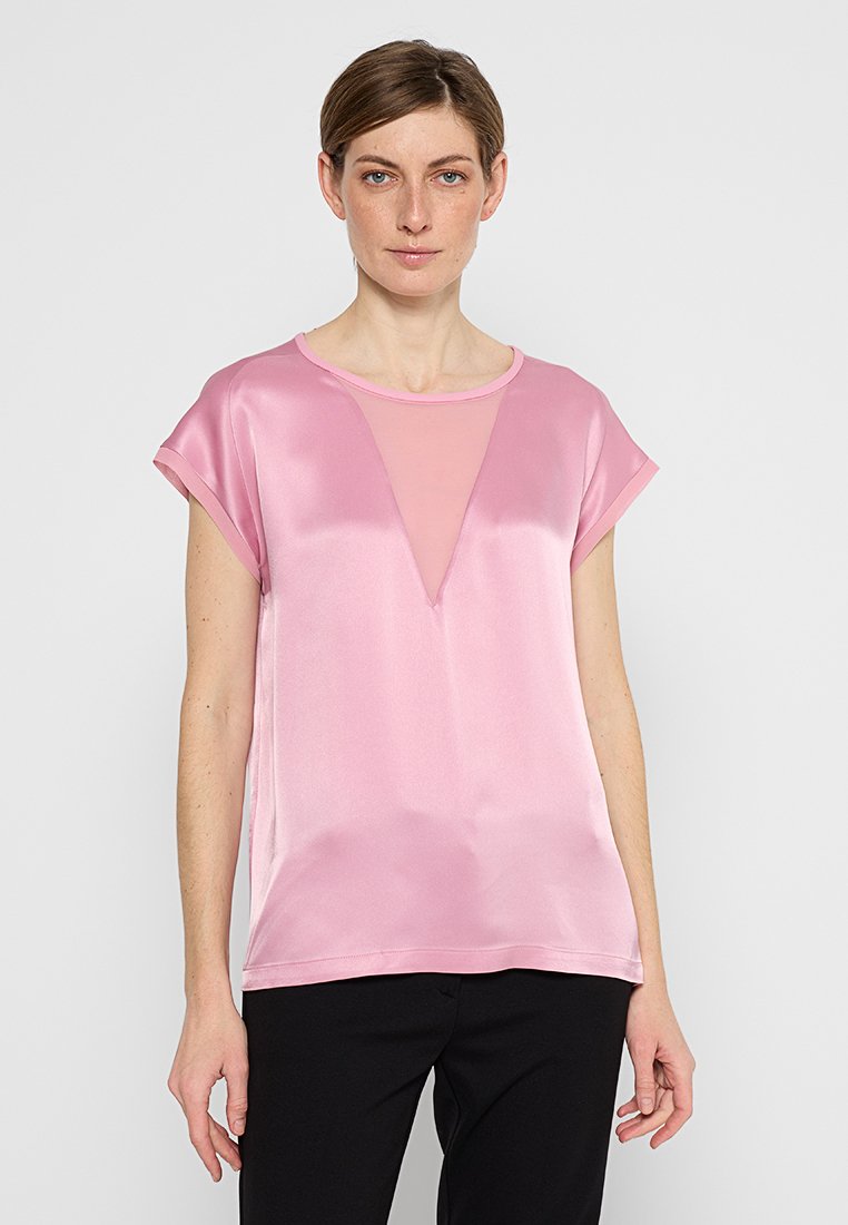 Eine Frau mit schulterlangen braunen Haaren trägt eine glänzende pinke Bluse mit kurzen Ärmeln und einem transparenten V-förmigen Einsatz sowie schwarze Hosen.