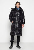 Mann steht in einem langen, glänzenden schwarzen Puffer-Mantel, blauer Rollkragenjacke, schwarzen Hosen und schwarzen Stiefeln vor einem schlichten weißen Hintergrund.