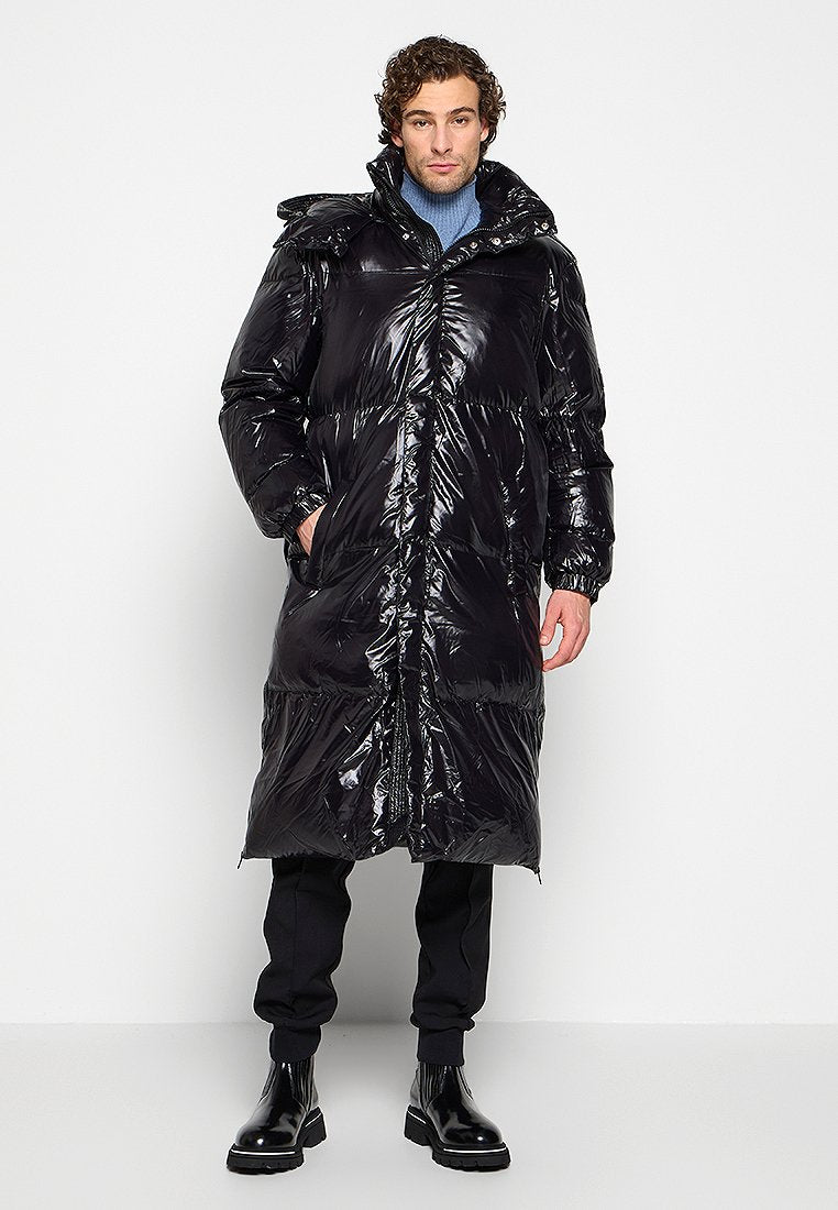 Mann steht in einem langen, glänzenden schwarzen Puffer-Mantel, blauer Rollkragenjacke, schwarzen Hosen und schwarzen Stiefeln vor einem schlichten weißen Hintergrund.