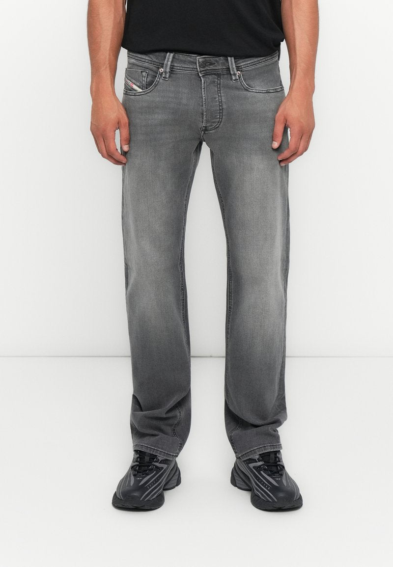 Mann in verwaschenen grauen, geraden Jeans, schwarzen Sneakers und einem schwarzen Shirt, stehend vor einem schlichten weißen Hintergrund.