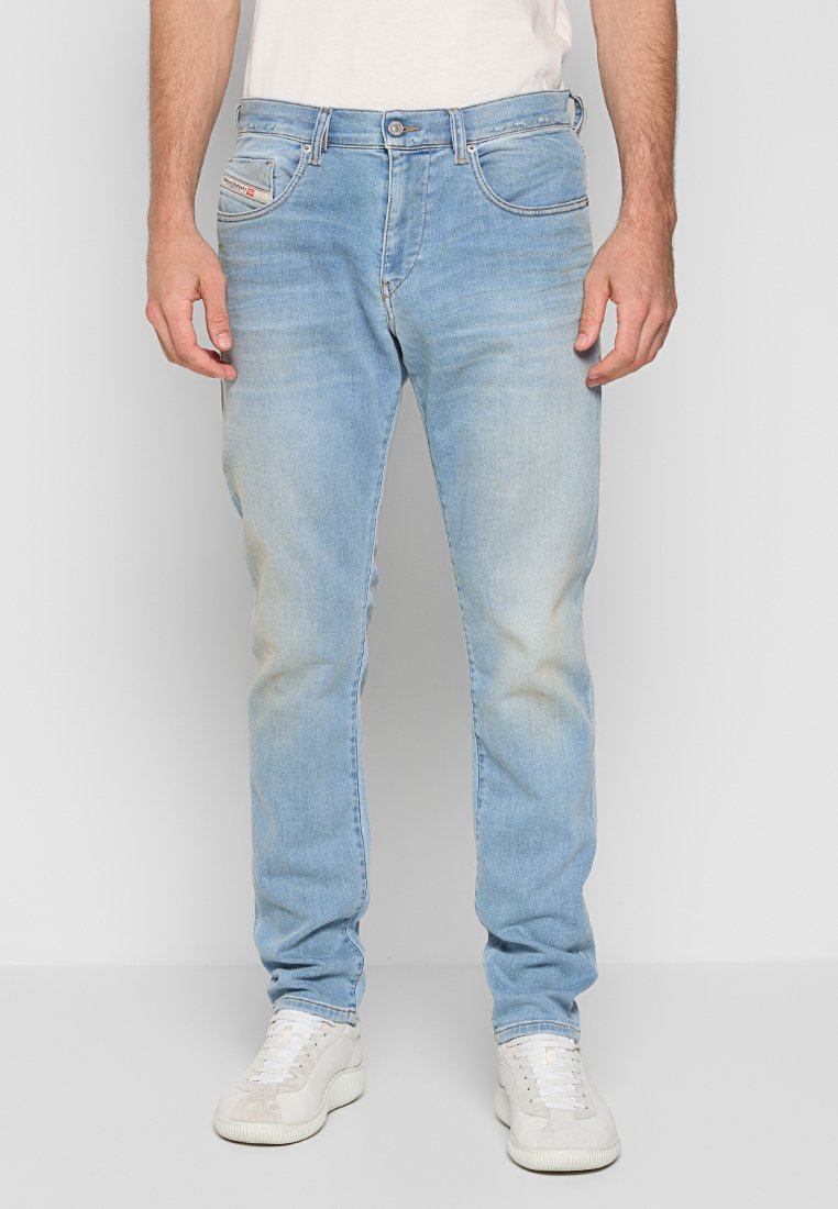 Mann in hellblauen, ausgebleichten Jeans und weißen Sneakers, der vor einem schlichten hellgrauen Hintergrund steht.