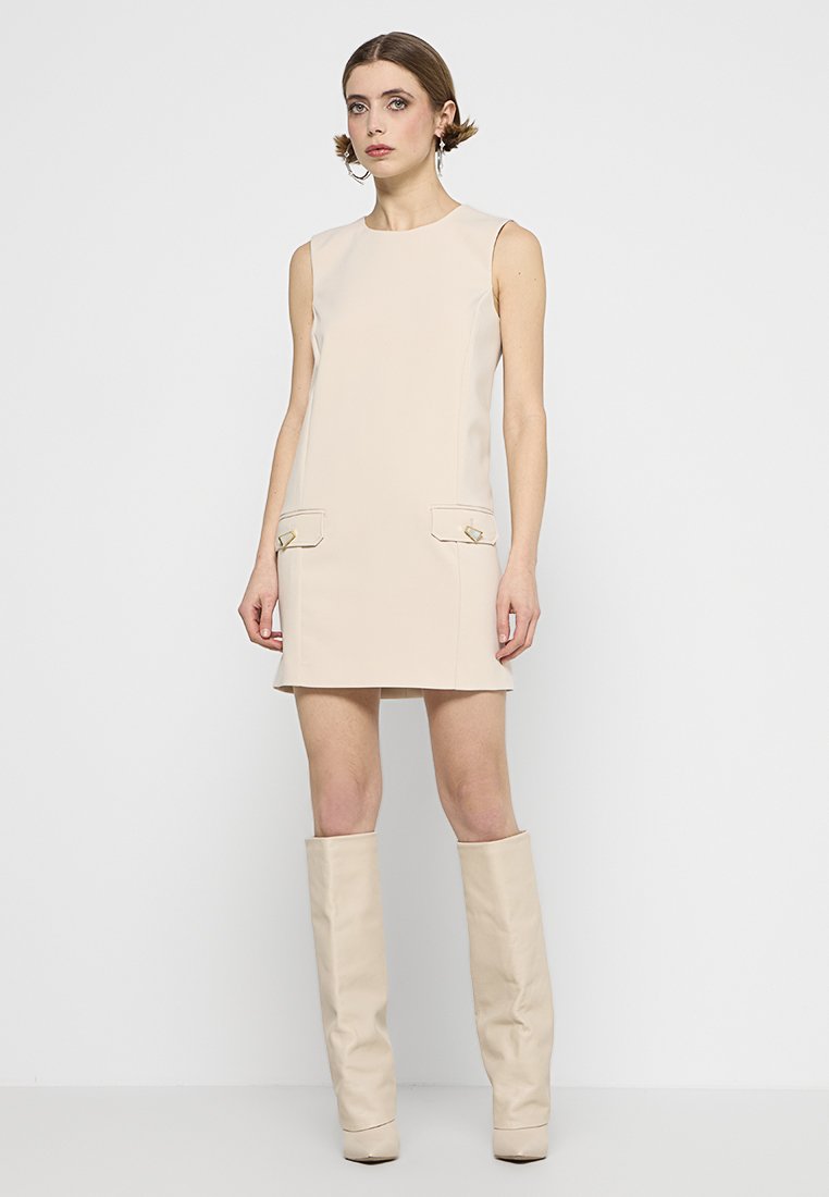 Beiges ärmelloses Kleid mit zwei vorderen Taschen, goldenen Akzenten, tailliertem Design und gerader Silhouette. Das Model trägt passende knielange Stiefel.