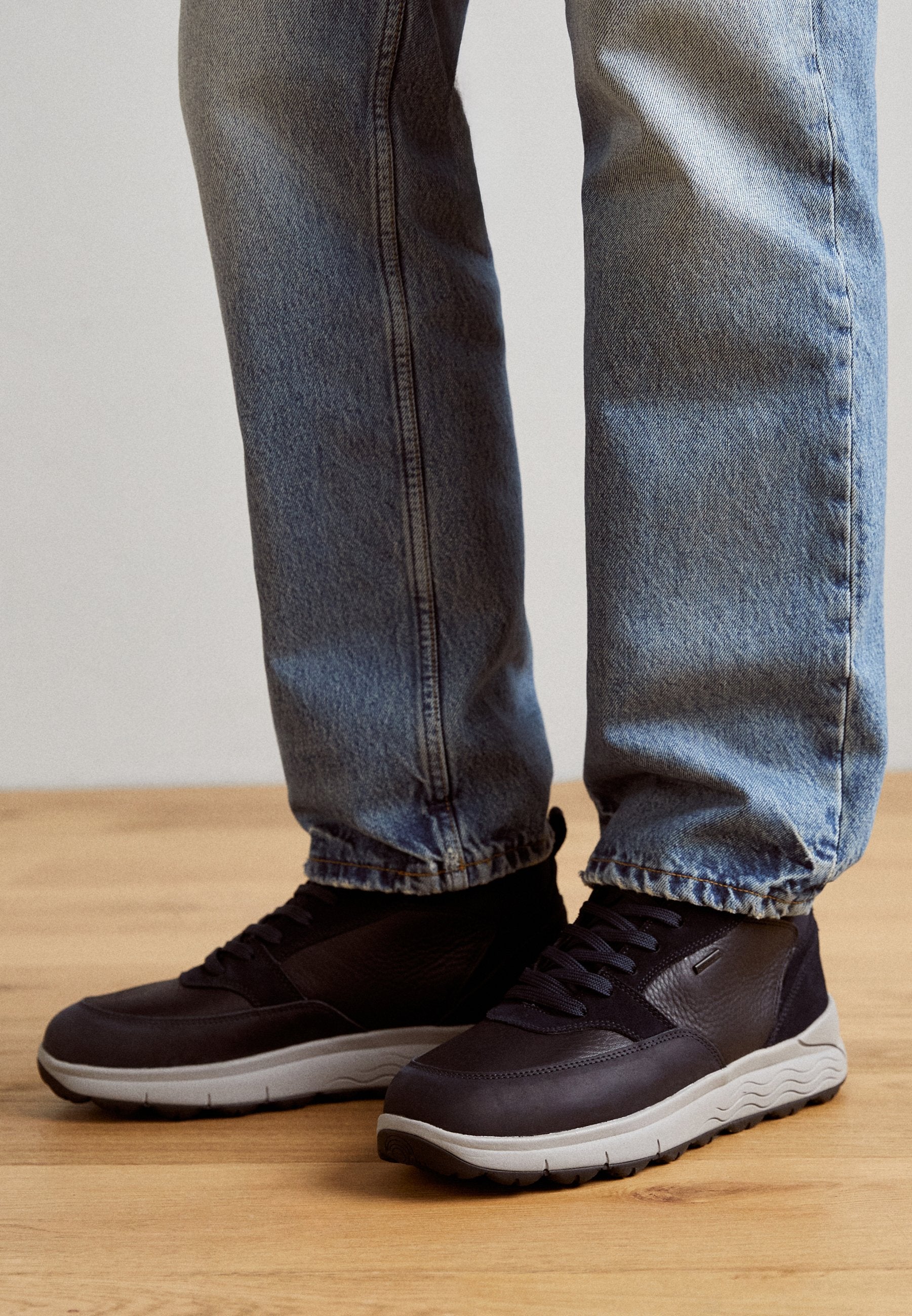 Schwarze Turnschuhe mit einem strukturierten Obermaterial aus Leder und einer grauen Gummisohle. Merkmale sind eine Schnürung und ein abgerundetes Design. Kombiniert mit blauen Jeans.