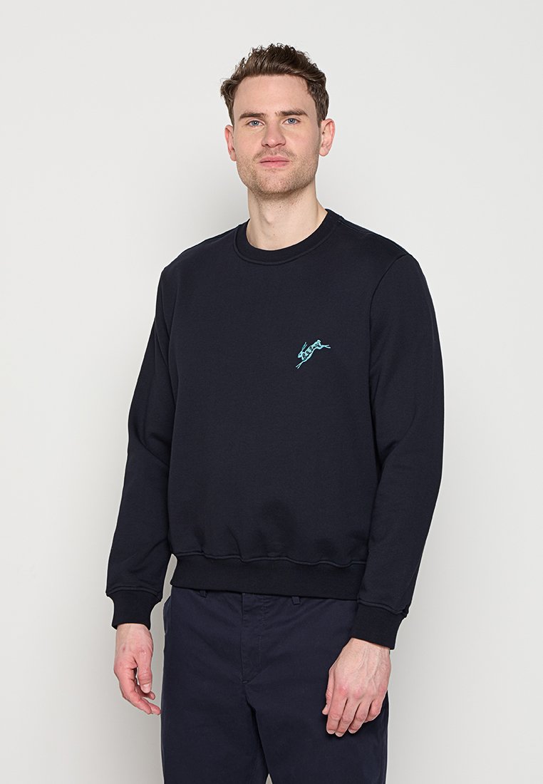 Mann trägt einen schlichten schwarzen Sweatshirt mit kleinem hellblauem rennendem Hundlogo auf der Brust, steht vor einem weißen Hintergrund.