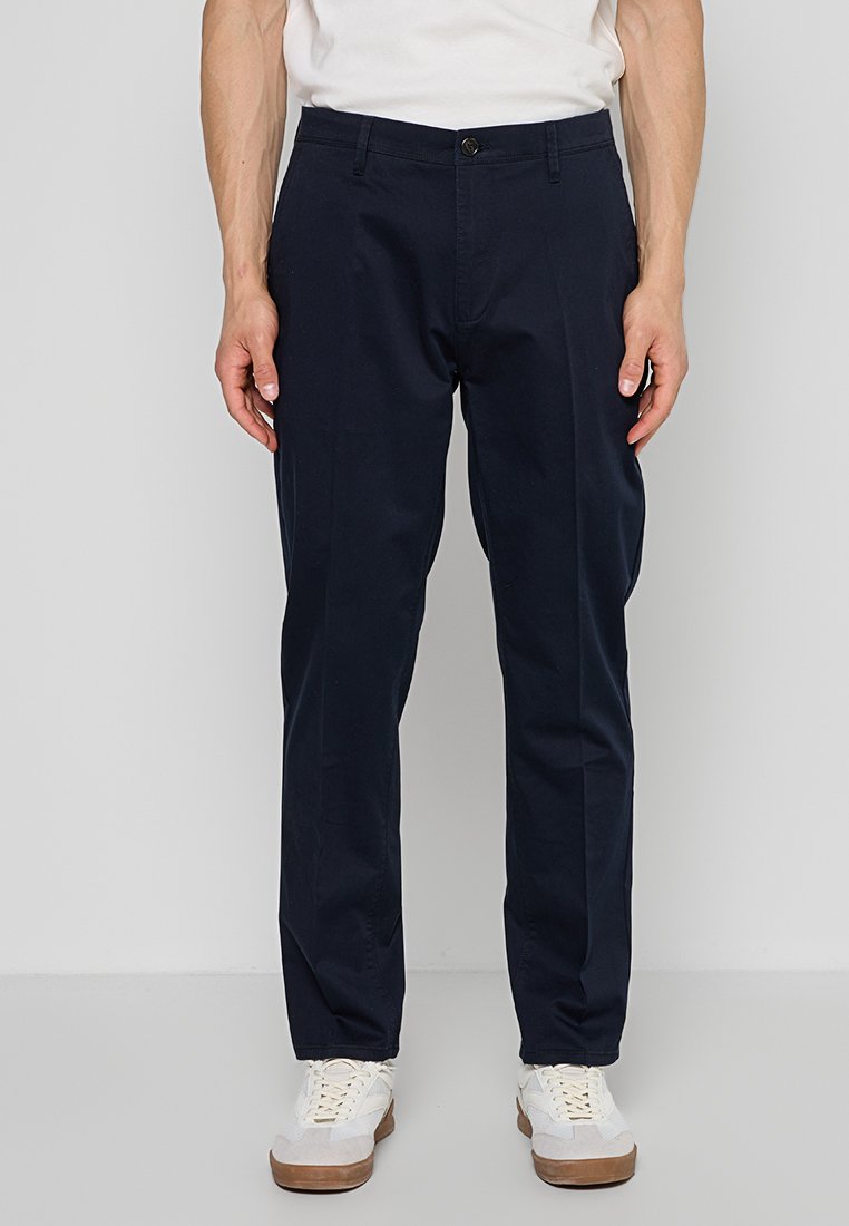 Mann trägt marineblaue gerade geschnittene Hose, weiße Sneakers und ein weißes Hemd, steht vor einem einfarbigen hellgrauen Hintergrund.