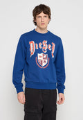 Junger Mann mit braunen Haaren, der einen blauen Diesel-Pullover mit rotem und weißem Wappen und schwarze Cargo-Hosen vor einfarbigem Hintergrund trägt.