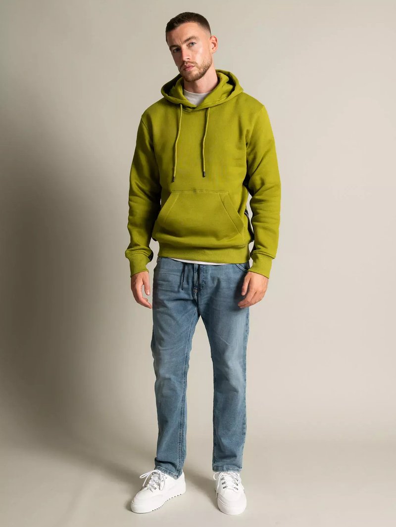 Grüne Kapuzenjacke mit Kordelzug, Kängurutasche, getragen über einem hellen Shirt. Kombiniert mit blauen Jeans und weißen Turnschuhen. Einfarbiger Hintergrund.