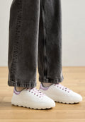 Weiße Sneaker mit glatter Textur, lila Akzenten und dicken, segmentierten Sohlen. Getragen mit lockeren, grauen Jeans.