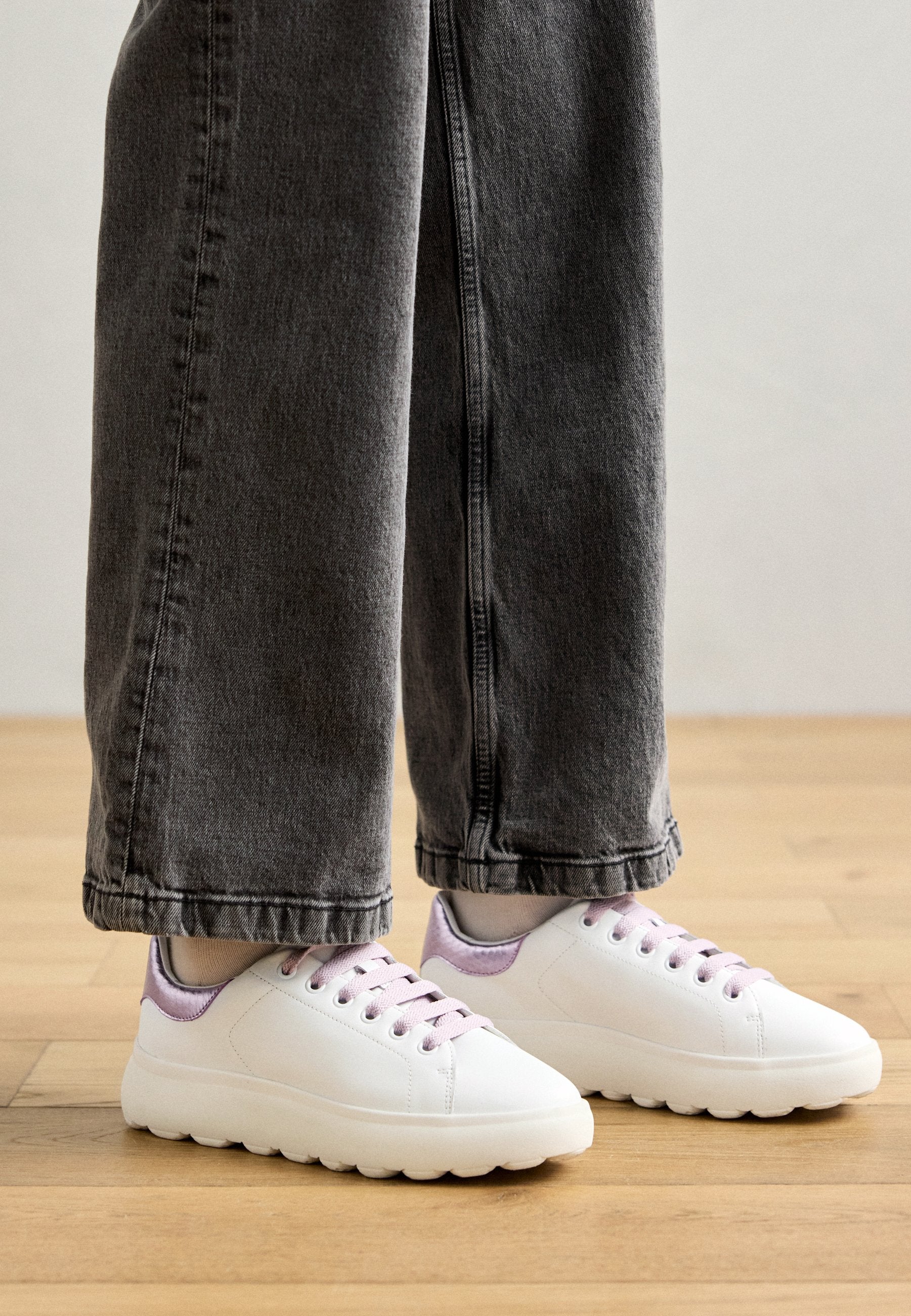 Weiße Sneaker mit glatter Textur, lila Akzenten und dicken, segmentierten Sohlen. Getragen mit lockeren, grauen Jeans.