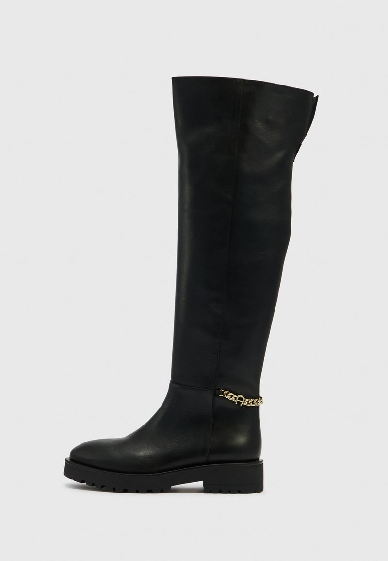 Schwarzer kniehoher Lederstiefel mit flacher Sohle, runder Zehenpartie und goldener Kettenverzierung am Knöchel. Glatte Textur mit minimaler Nahtführung.