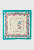 Quadratischer cremefarbener Schal mit türkisfarbenem Rand, rot-blauem Blumenmuster an den Rändern und zentralen stilisierten Buchstaben, die ein kreisförmiges Design bilden.