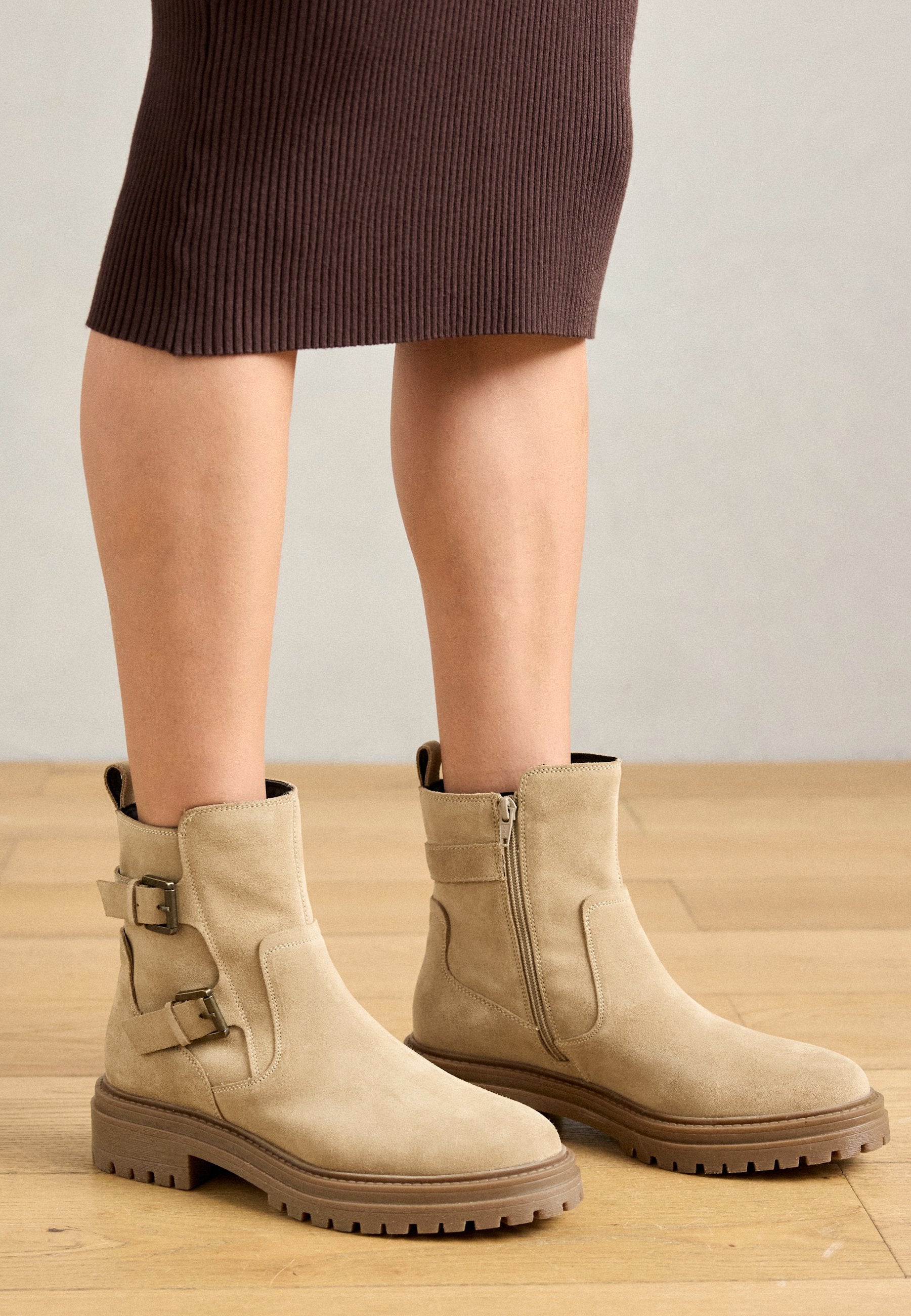Beige Wildleder Stiefeletten mit zwei seitlichen Schnallen und einem Reißverschluss. Dicke braune Gummisohlen mit geriffelter Textur für besseren Halt.