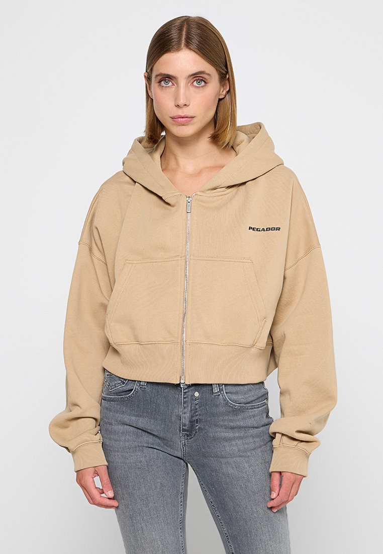 Beige Crop-Hoodie aus weichem Stoff mit Reißverschluss, Fronttaschen und schwarzem "PEGADOR"-Logo auf der linken Brust.