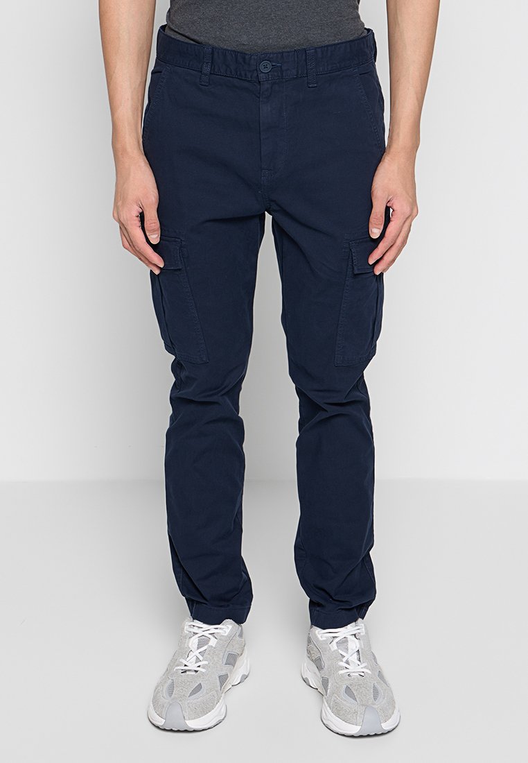 Navyblaue Cargo-Hosen aus Baumwolle. Zu den Merkmalen gehören mehrere Seitentaschen und eine schmal zulaufende Passform. Getragen mit grauen Turnschuhen.