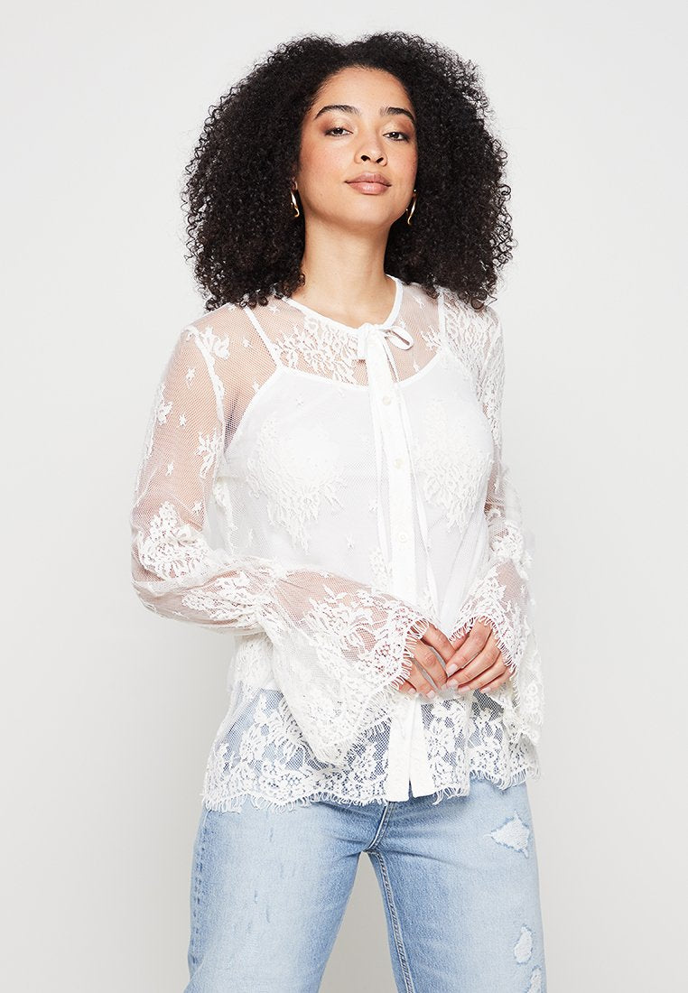 Frau mit lockigem Haar, die eine weiße Spitzenbluse mit langen Ärmeln und floralen Mustern sowie hellblauen, verwaschenen Jeans trägt, steht mit gefalteten Händen.