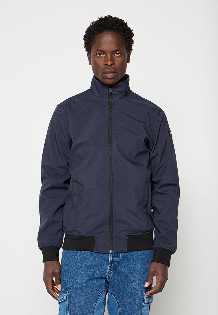Mann trägt eine dunkelblaue Jacke mit Reißverschluss, schwarzen Bündchen und Bund, steht mit entspannten Händen seitlich vor einfarbigem Hintergrund.