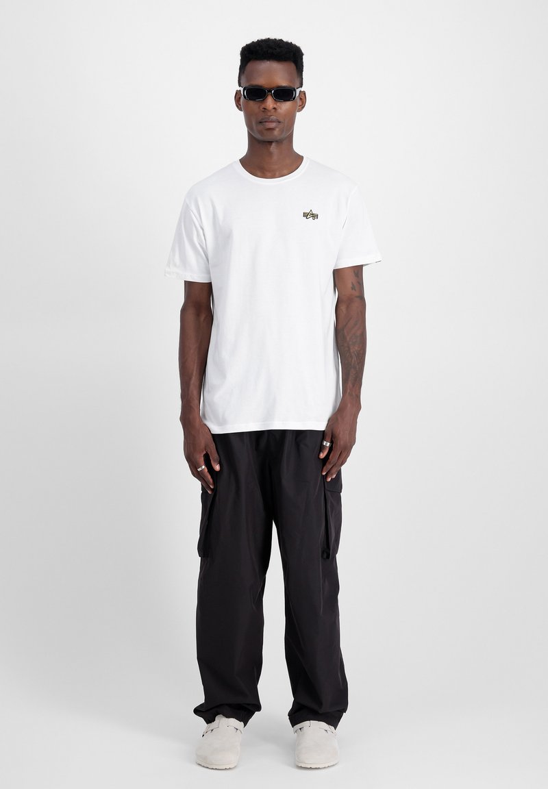 Weißes Baumwoll-T-Shirt mit kleinem grünen und goldenen Logo, kombiniert mit weiten schwarzen Cargo-Hosen und weißen Schuhen. Model trägt Sonnenbrille.